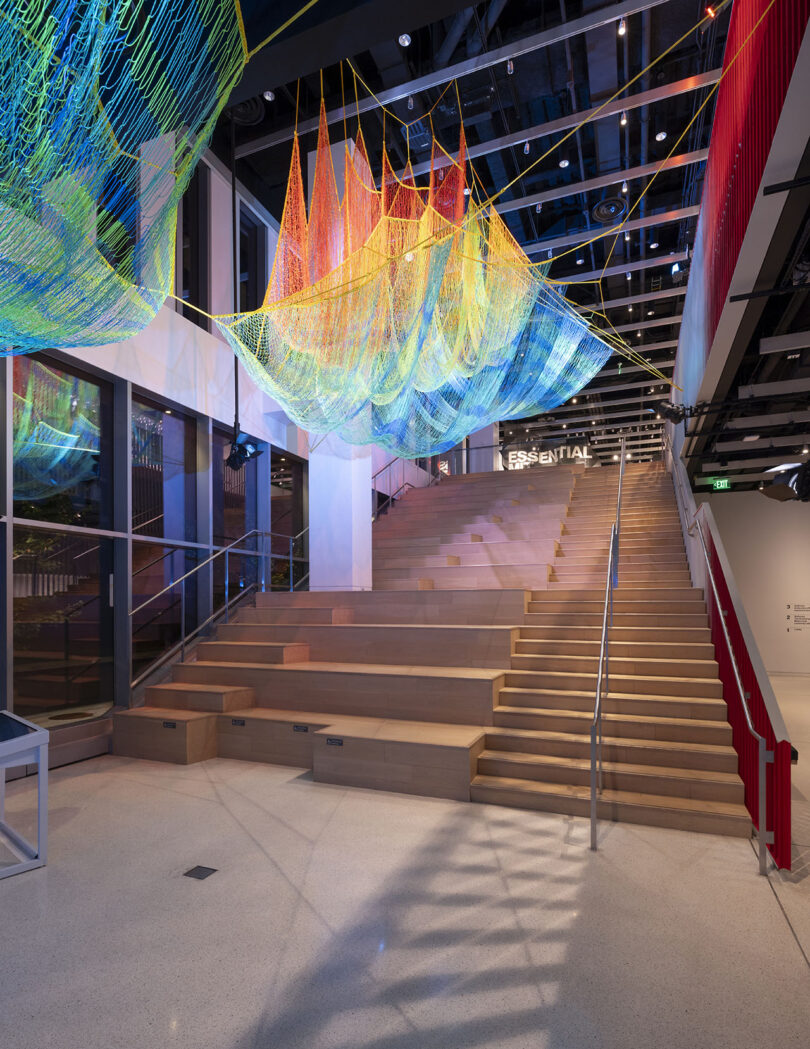 A wide wooden staircase with metal railings features a colorful net installation suspended above in a modern interior space with high ceilings and large windows.