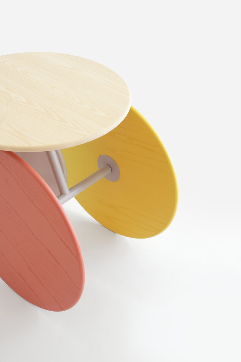 A round light wood tabletop supported by three angled oval legs in pink, yellow, and natural wood colors on a white background.