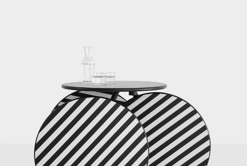 A small round table with bold black and white diagonal stripes holds a clear glass carafe and a glass of water against a plain light background.