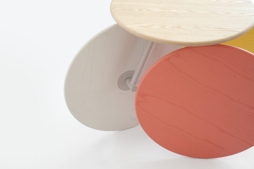 Three round wooden tabletops in light wood, white, and red colors overlap slightly, connected by a central support on a white background.