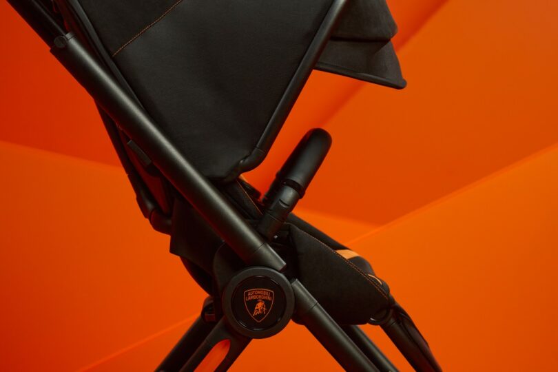 Close-up of a Silver Cross Lamborghini black stroller handle with stitching detail against an orange background.