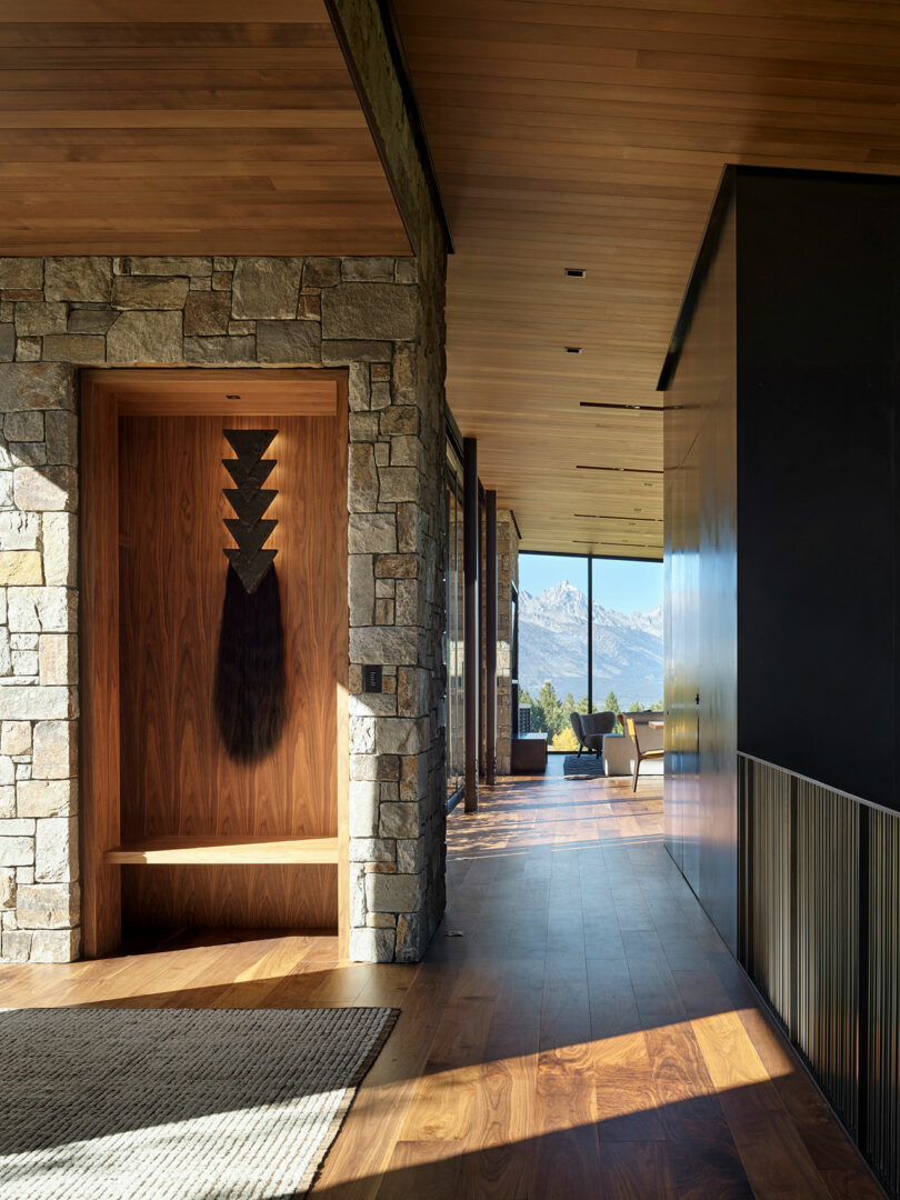 A modern hallway with stone and wood walls, wooden floors, a bench with wall art, and large windows overlooking mountains in the background.