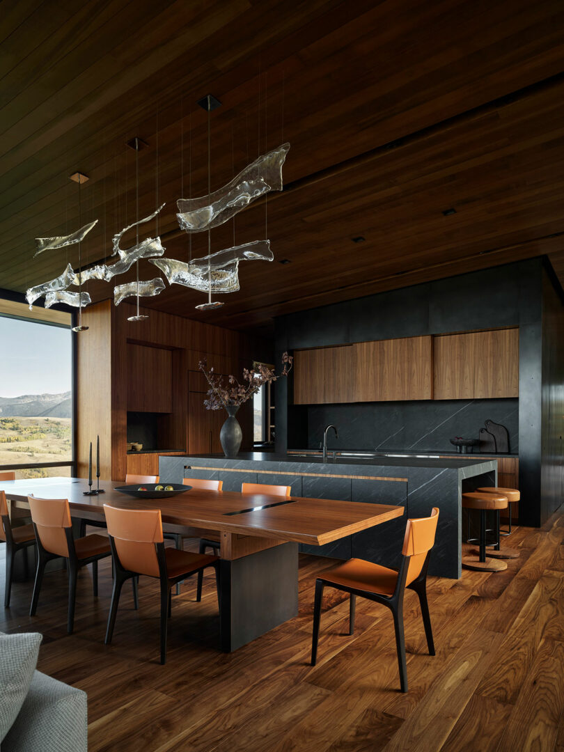 Modern kitchen and dining area with wood floors, wood-paneled walls and ceiling, black countertops, tan chairs, and abstract glass pendant lights, with large windows showing an outdoor view.