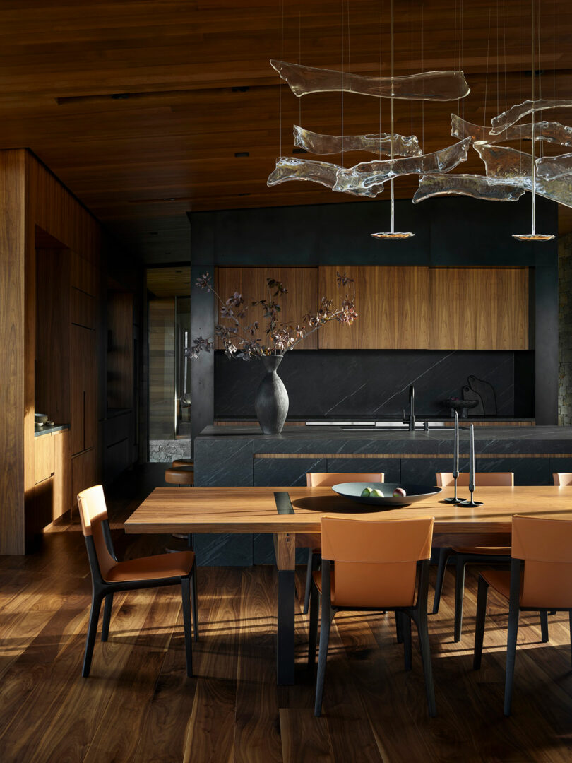 Modern kitchen and dining area with wooden floors, cabinets, and table, orange chairs, dark stone countertops, and abstract glass pendant lights above.