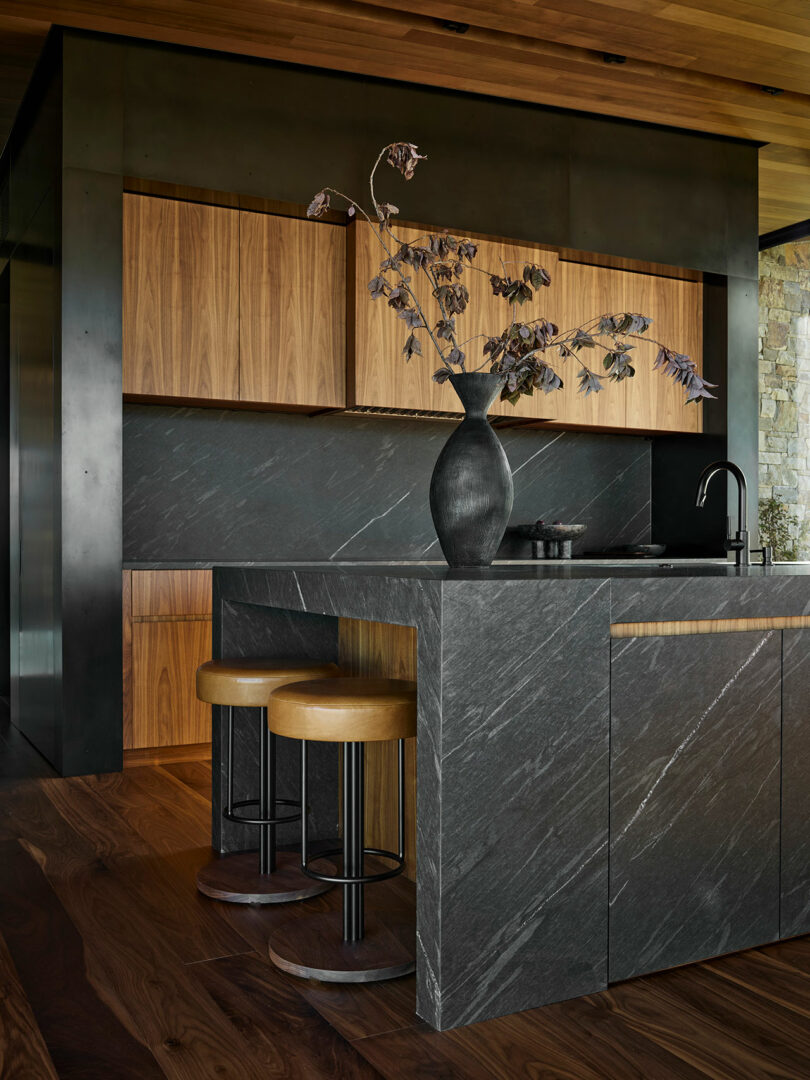 Modern kitchen with dark stone countertops and island, wood cabinetry, two stools, and a black vase with dried branches on the island.