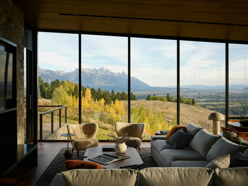 Modern living room with large glass windows offering a scenic view of mountains, trees, and an open landscape. Contemporary furniture and décor are arranged inside.