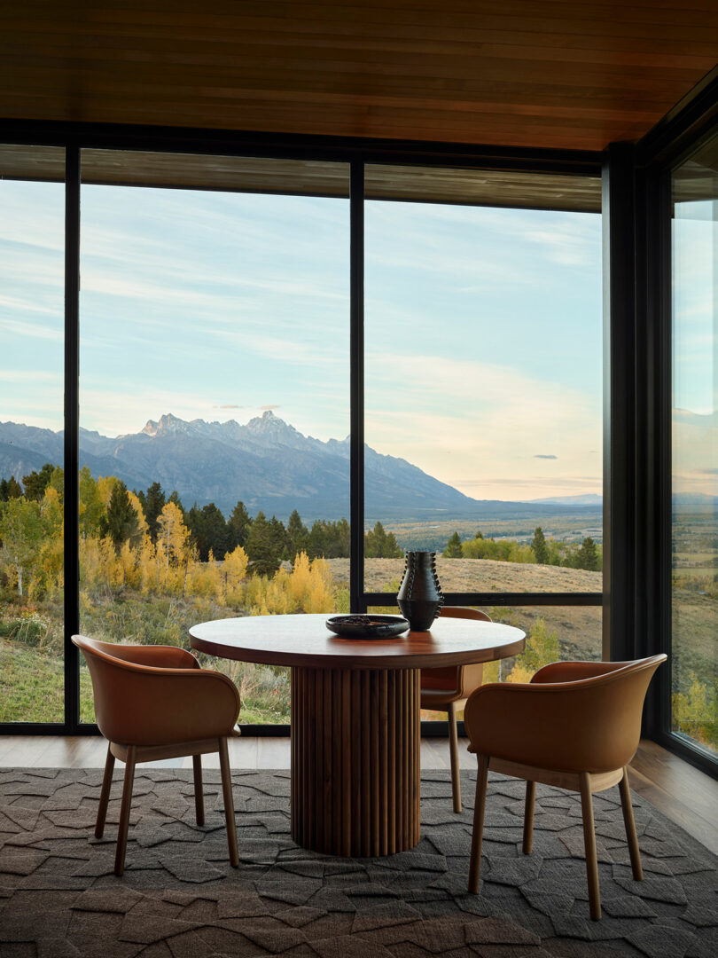 A round wooden table with two chairs sits by large floor-to-ceiling windows overlooking a scenic landscape of mountains, trees, and sky.