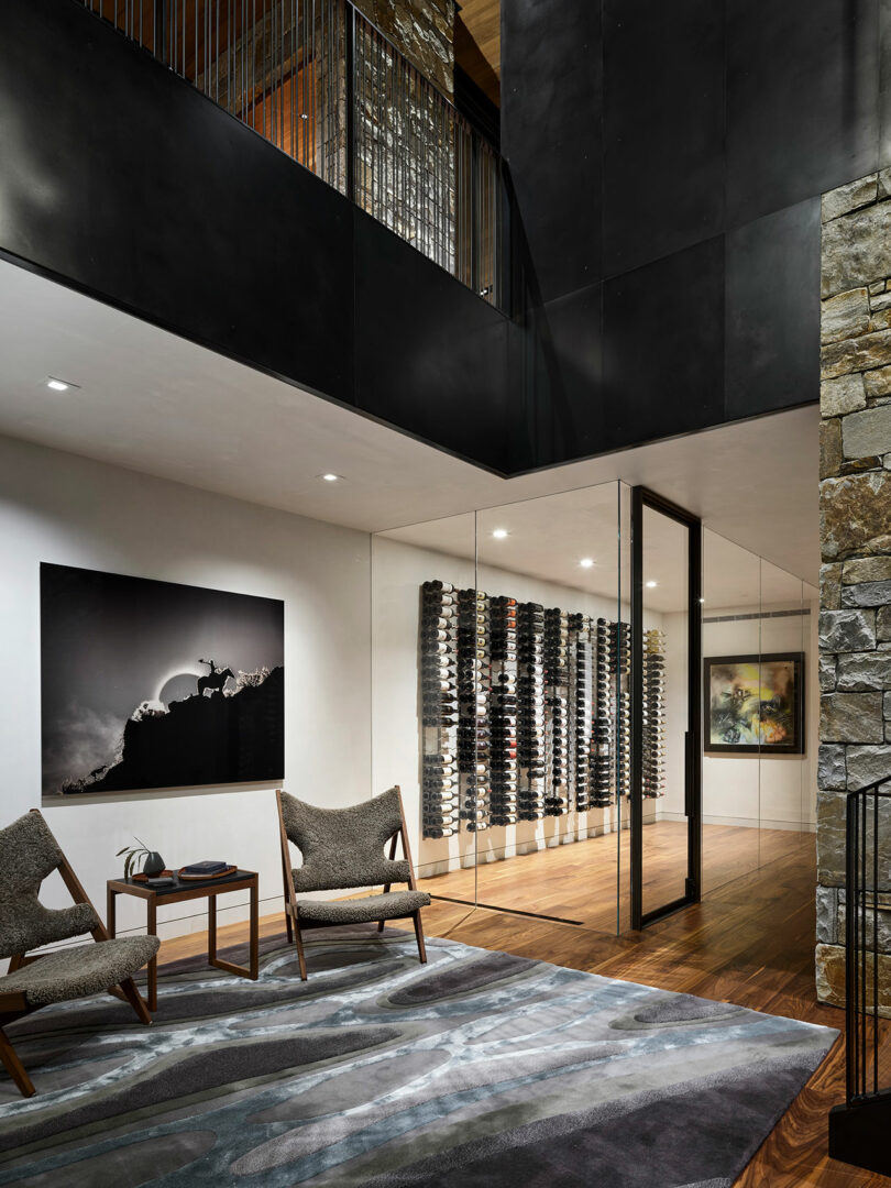 Modern interior with two chairs, a small table, a wine rack behind glass doors, stone and metal walls, artwork, and a patterned rug on a wooden floor.