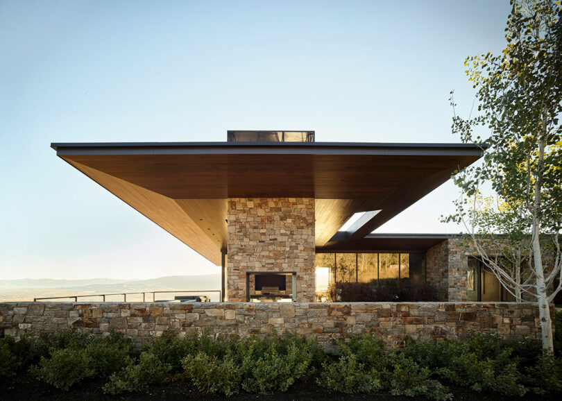Modern house with wide, overhanging wood roof, stone walls, large windows, and an outdoor fireplace, set against a natural landscape and clear sky.