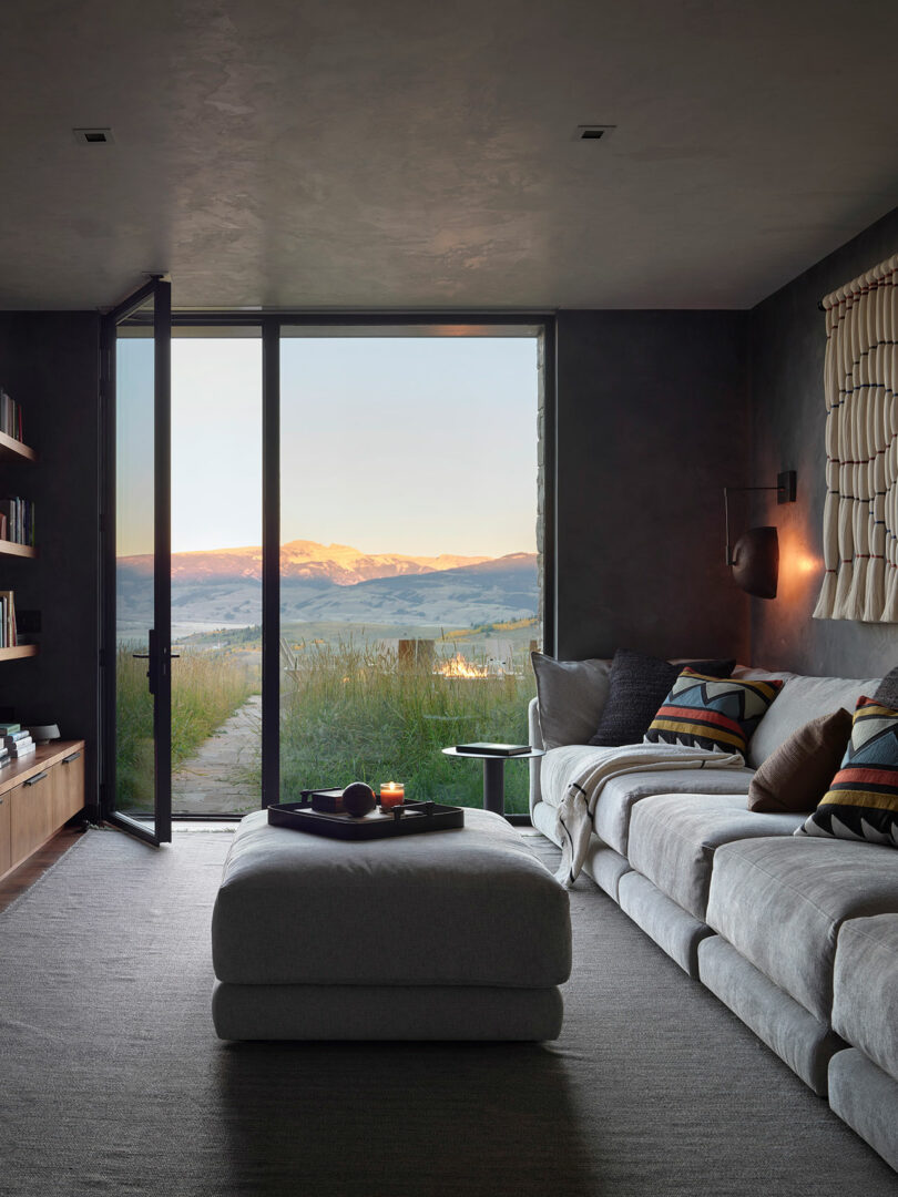 A modern living room with a gray sectional sofa, large ottoman, and open glass door revealing a scenic mountain landscape at sunset.