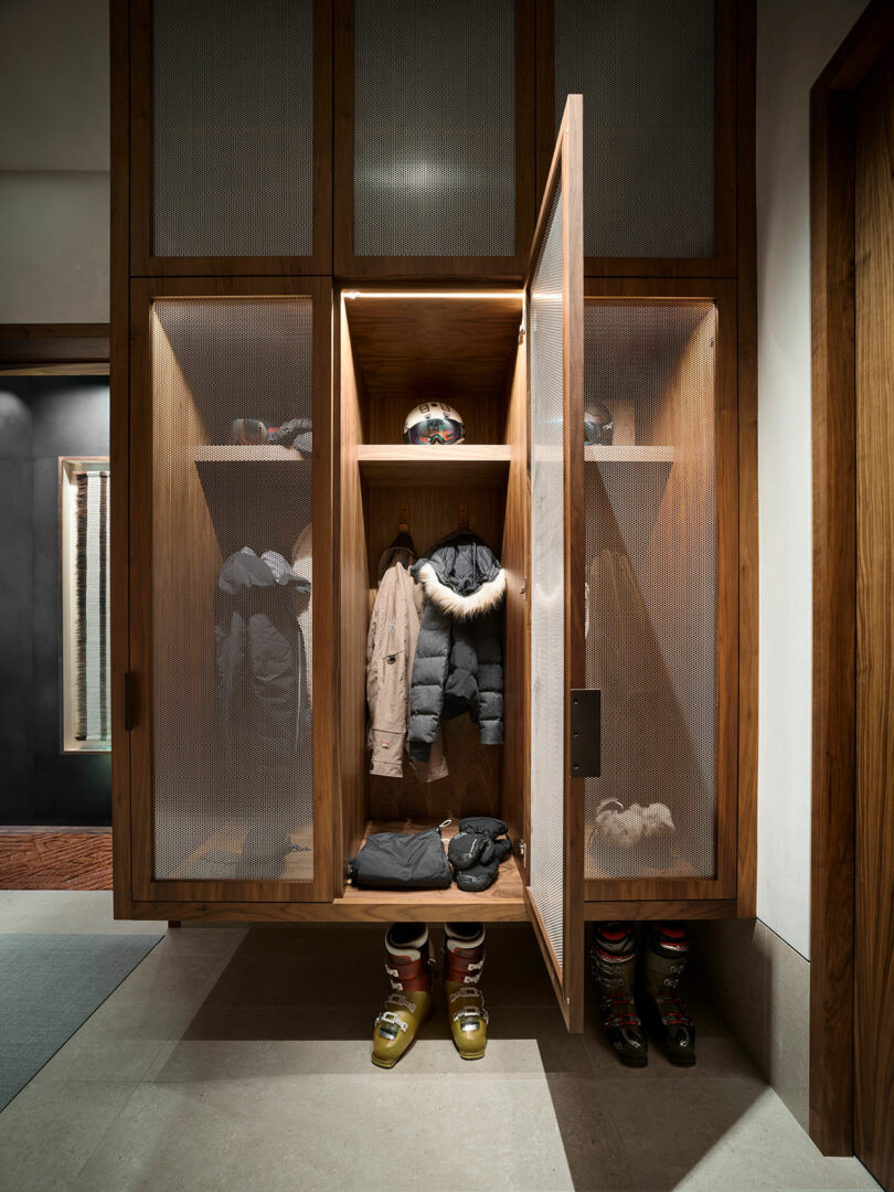 Wooden locker with mesh doors, containing winter coats, helmets, gloves, and ski boots neatly arranged on shelves and the floor.