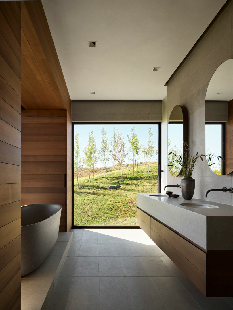 A modern bathroom with wood and stone finishes, a freestanding tub, a large vanity, and a floor-to-ceiling window overlooking a grassy outdoor landscape.