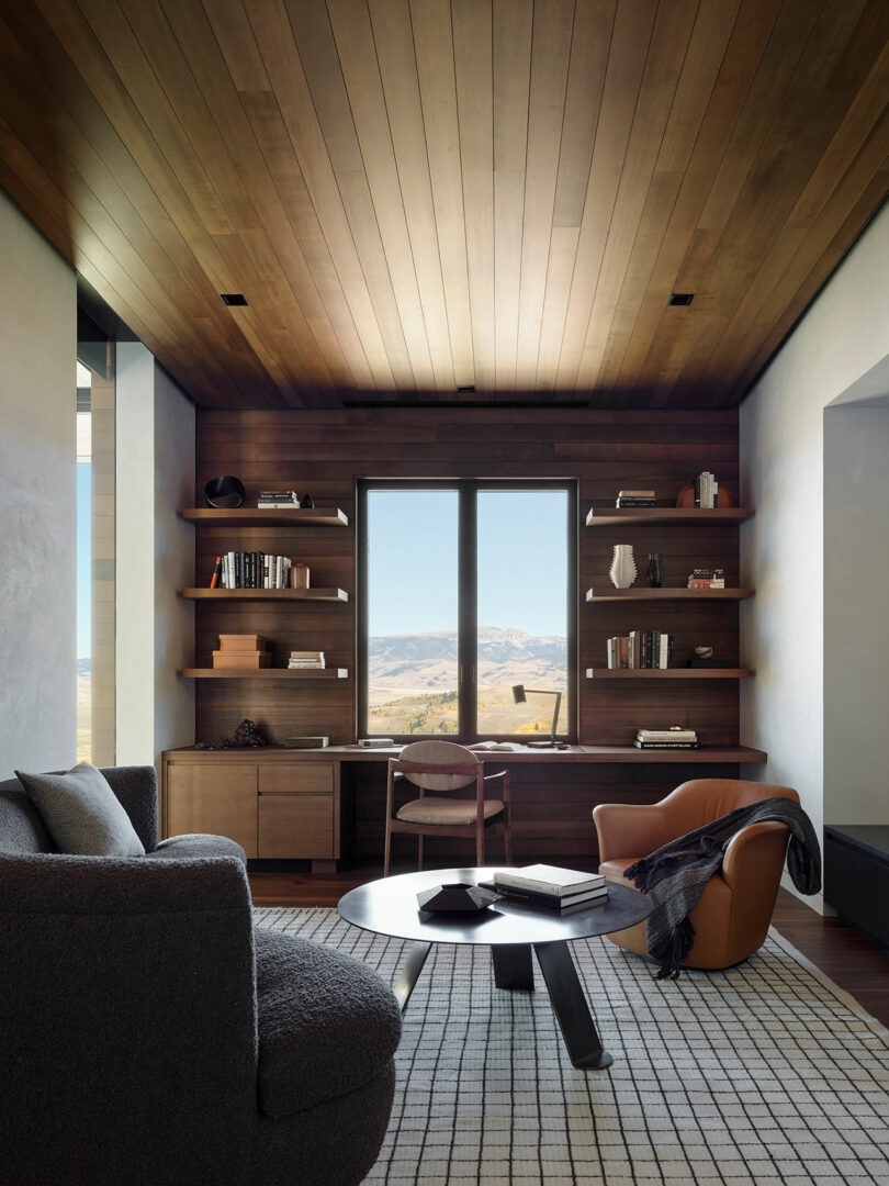 Modern home office with wooden ceiling and shelves, a desk facing a window with a mountain view, a gray sofa, a brown armchair, and a round coffee table on a patterned rug.