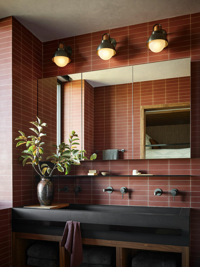 Modern bathroom with red tile walls, a black countertop, round sink, a large mirror, three wall-mounted lights, and a vase with branches on the counter.