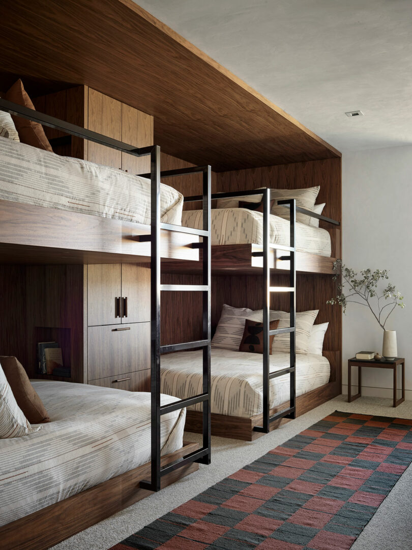 Four wooden bunk beds with metal ladders are arranged against a wall in a modern room; a side table with a vase and a geometric rug are also visible.