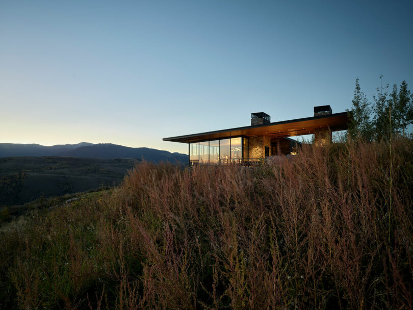 Modern house with large glass windows situated on a grassy hillside at dusk, overlooking distant mountains under a clear sky.