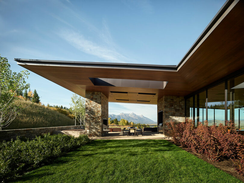 A modern house with a wide overhanging roof, stone and glass walls, a covered outdoor patio, and landscaped lawn, with mountains visible in the background.