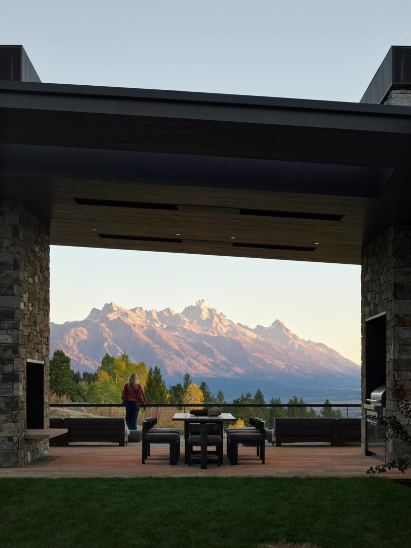 A person sits on a patio under a covered area, facing outdoor furniture and a scenic view of snow-capped mountains at sunset.