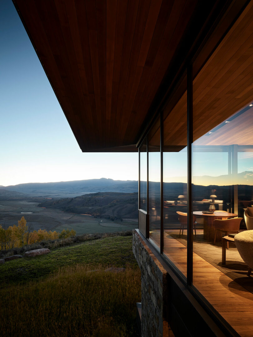Modern house with large glass windows overlooks a scenic valley and distant mountains at sunset, with warm interior lighting visible inside.