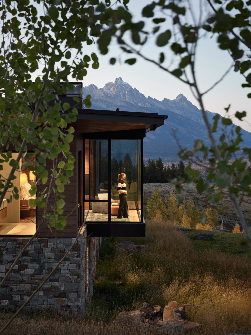 A person stands by the window of a modern house with glass walls, overlooking a grassy landscape and mountains at dusk.