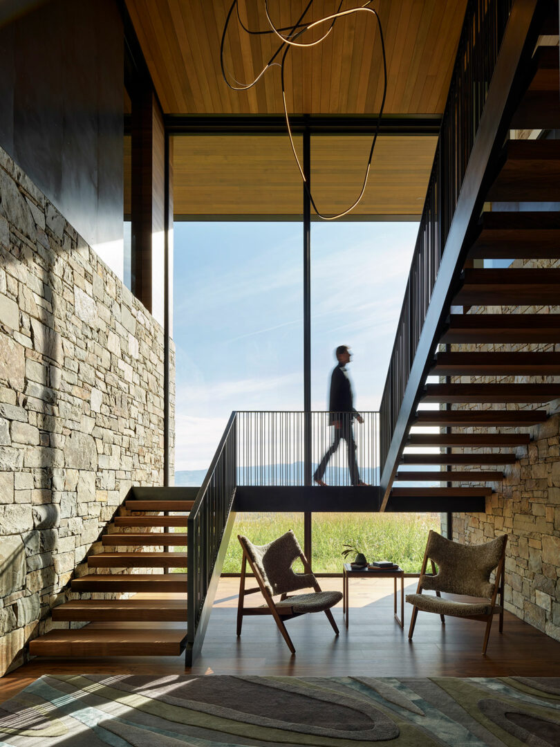 A person walks on an upper-level walkway in a modern interior with stone walls, large windows, and mid-century chairs near a coffee table on the lower level.