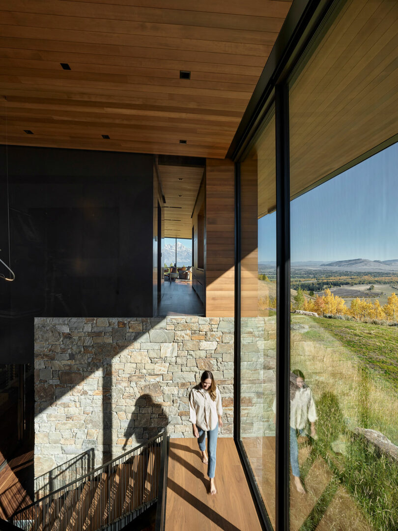 A woman walks up a wooden staircase inside a modern house with large glass windows overlooking a scenic, grassy landscape and distant mountains.
