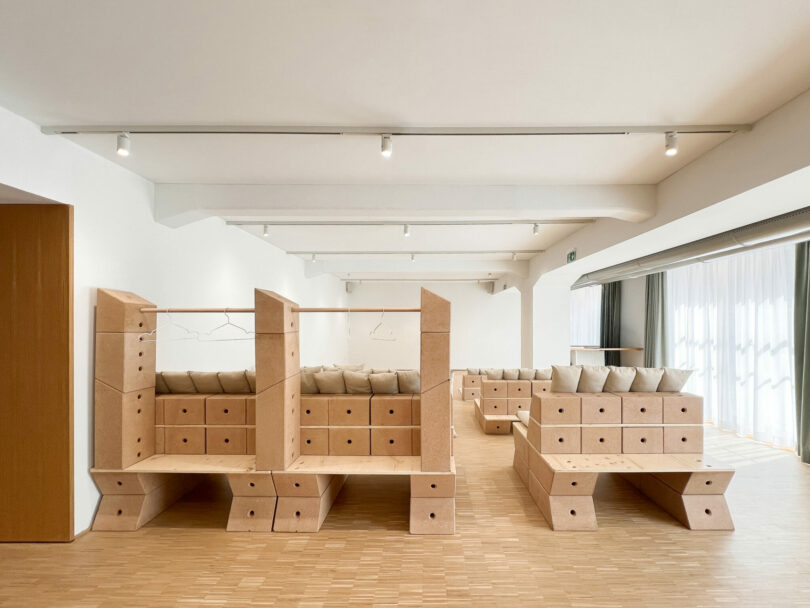 Minimalist room with light wood floors, white walls, and modular wooden seating units with beige cushions arranged in rows. Ceiling lights and large windows with sheer curtains in background