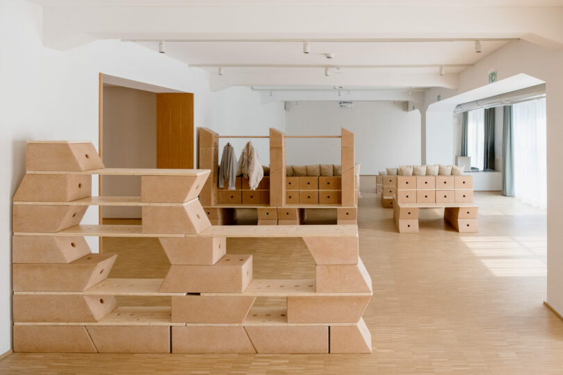 A modern, minimalist room with modular wooden shelving and seating units, light wood flooring, white walls, and coats hanging on the racks