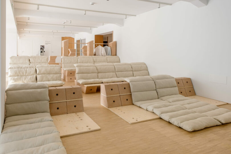 A room with rows of low, padded beige lounge seats on wooden platforms, arranged in a stepped formation, with white walls and minimal decor