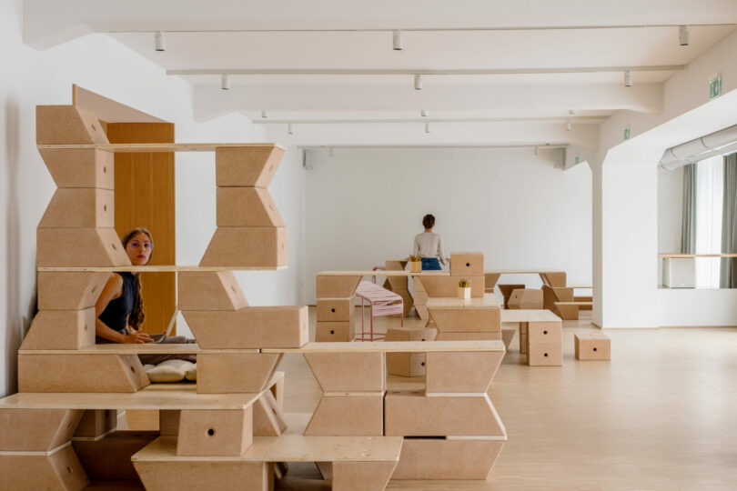 Two people are in a minimalist room with geometric wooden furniture; one woman sits behind a hexagonal shelf, while another stands by a table in the background