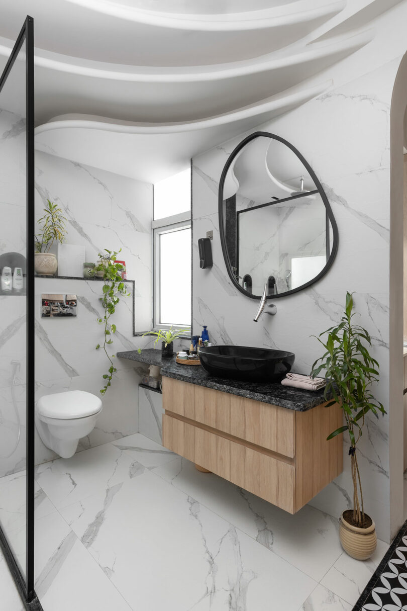Modern bathroom with white marble walls and floor, floating wood vanity with black sink, round mirror, glass shower partition, potted plants, and a wall-mounted toilet.