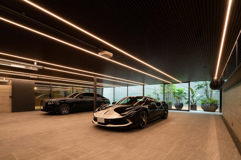 A modern garage with two luxury cars parked inside—one black sedan and one black-and-white sports car—under linear ceiling lights, with large windows showing plants outside.