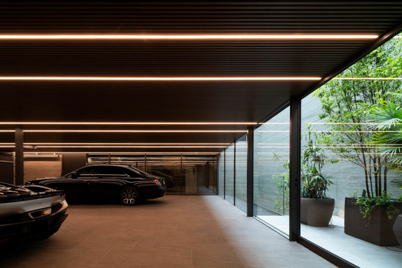 Modern indoor garage with two luxury cars, linear ceiling lights, large glass windows, and potted plants along the right side.