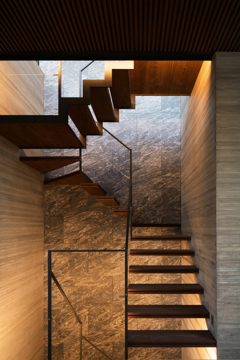 A modern staircase with dark wood steps and metal railings, set against marble-patterned walls and illuminated by warm recessed lighting.