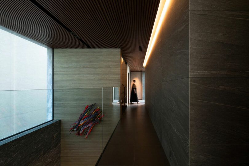 A modern hallway with stone walls, a slatted wood ceiling, abstract wall art, and a blurred figure walking in the distance.