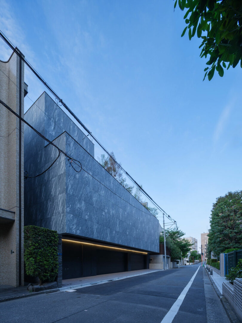Modern building with a dark stone facade and garage doors facing a quiet, empty street lined with greenery under a clear blue sky.