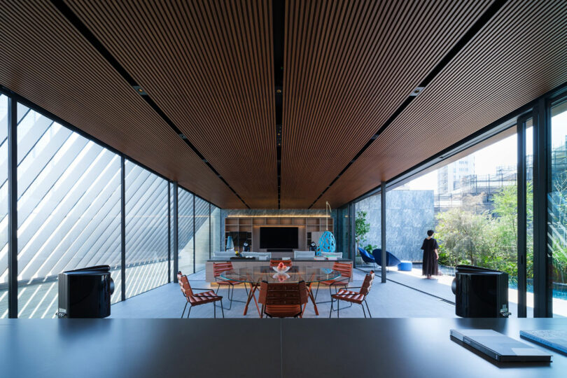 Modern living room with floor-to-ceiling windows, wood slat ceiling, contemporary furniture, and a view of a person standing outside on a patio near greenery.