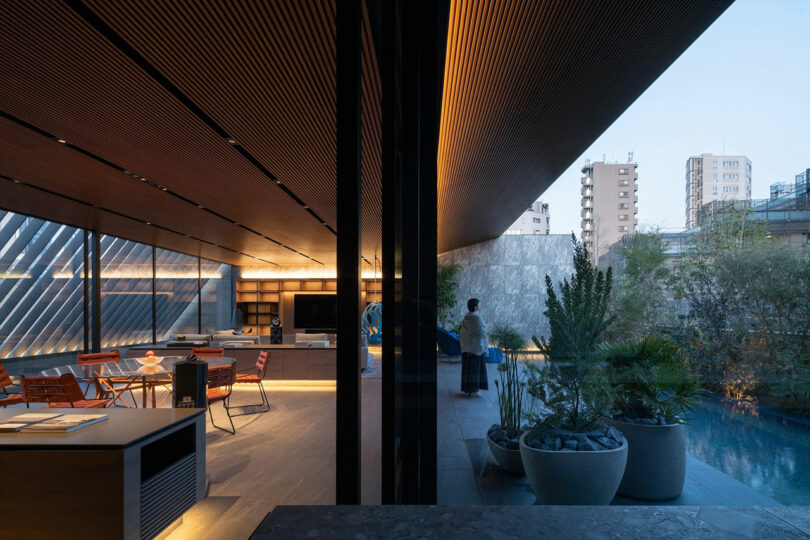 Modern indoor living space with wooden ceiling, dining table, and open view to an outdoor pool area with plants; a person stands near the pool at dusk.