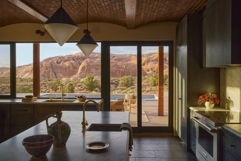 Modern kitchen with dark cabinets and pendant lights, large windows and doors open to a patio with a pool and scenic view of red rock desert cliffs.