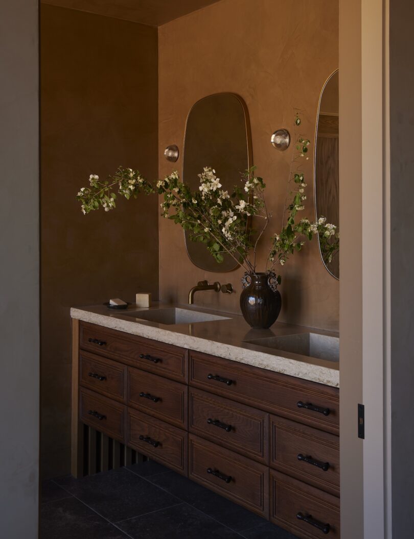 A bathroom vanity with a marble countertop, dual sinks, wooden drawers, two mirrors, a dark vase with branches, and warm brown walls.