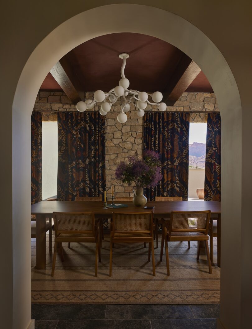 A dining room with a wooden table, six chairs, a modern white chandelier, stone walls, patterned curtains, and a vase of flowers, viewed through an arched doorway.