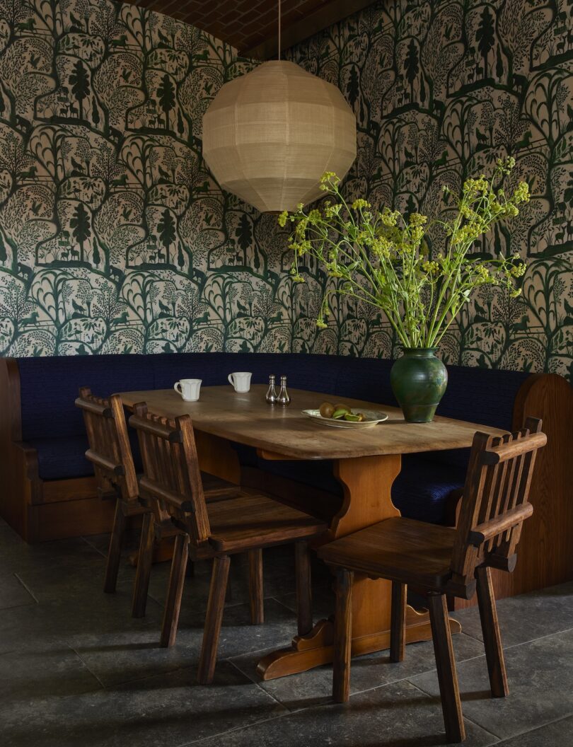 A wooden dining table with four chairs sits next to a built-in bench, with a large vase of green flowers and two cups; wallpaper with an animal motif decorates the walls.