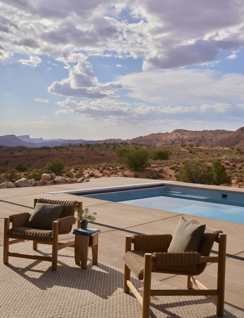 Two wicker chairs with cushions and a small table sit by a swimming pool, overlooking a desert landscape with hills and a cloudy sky in the background.
