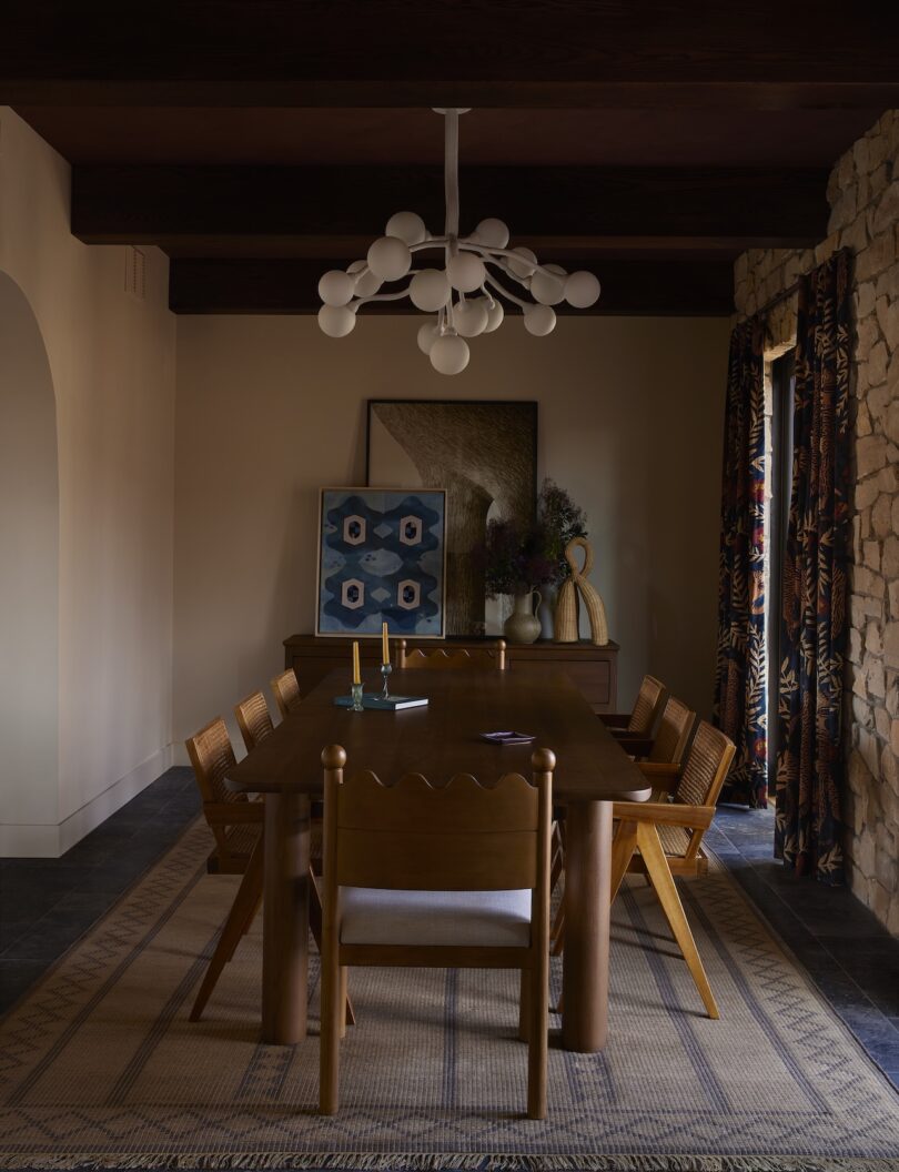 A dining room with a wooden table and six chairs, a modern chandelier, a sideboard with decor, framed art, and patterned curtains, featuring neutral and earthy tones.