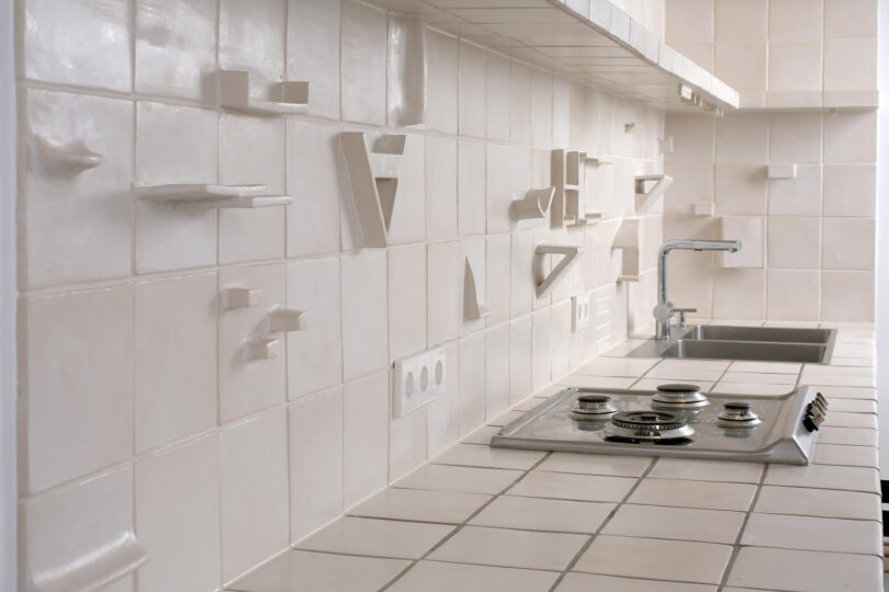 A modern kitchen with white tiled surfaces and backsplash, featuring abstract, geometric tile protrusions, a sink, a stovetop, and power outlets.