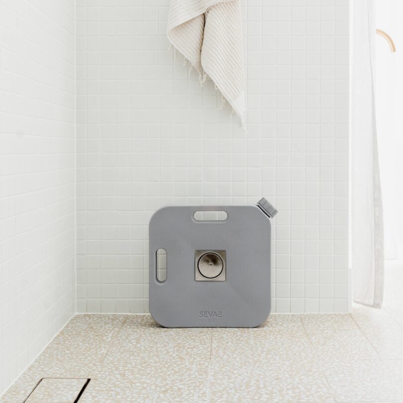 A square-shaped gray water container by SEVAS with a spout and handle sits on a tiled bathroom floor against a white tiled wall; a towel hangs above it.