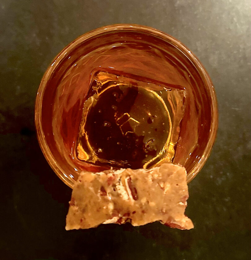 A glass of amber-colored liquor with a large ice cube, topped with a piece of nut-studded brittle, viewed from above.