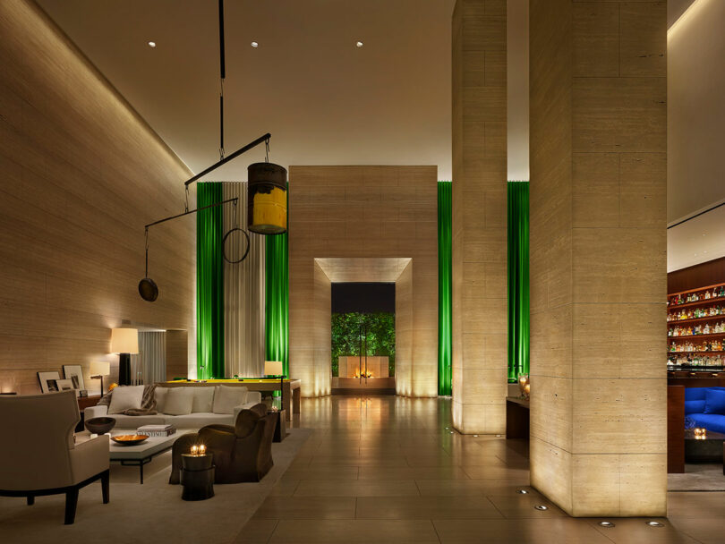 Modern West Hollywood EDITION hotel lobby with high ceilings, beige stone walls, tall green-lit columns, contemporary furniture, and a well-lit bar area on the right.