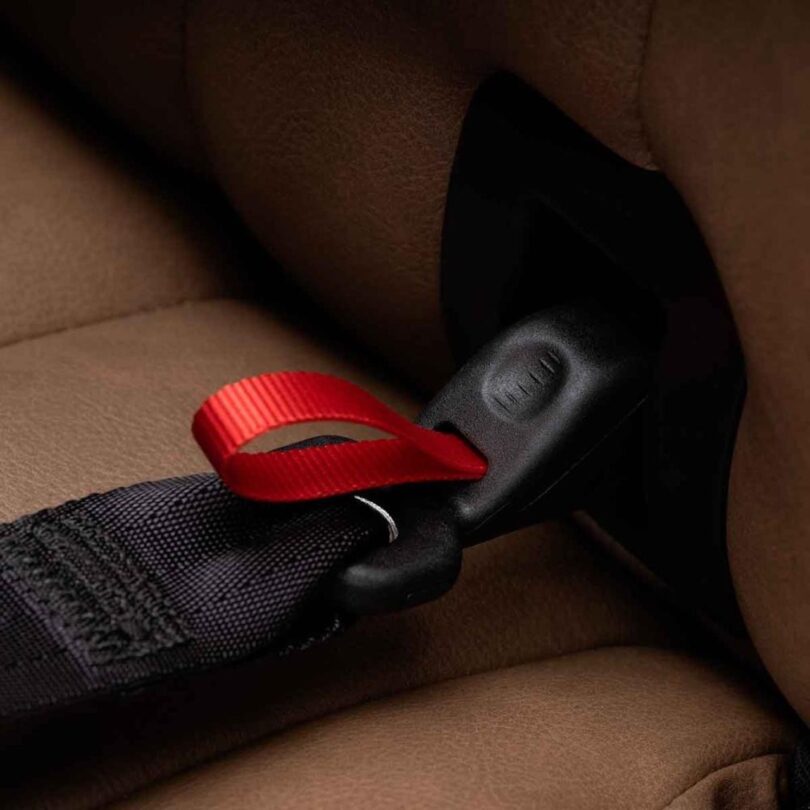 Close-up of a car seatbelt buckle with a black strap and a red pull tab, set against a tan seat background