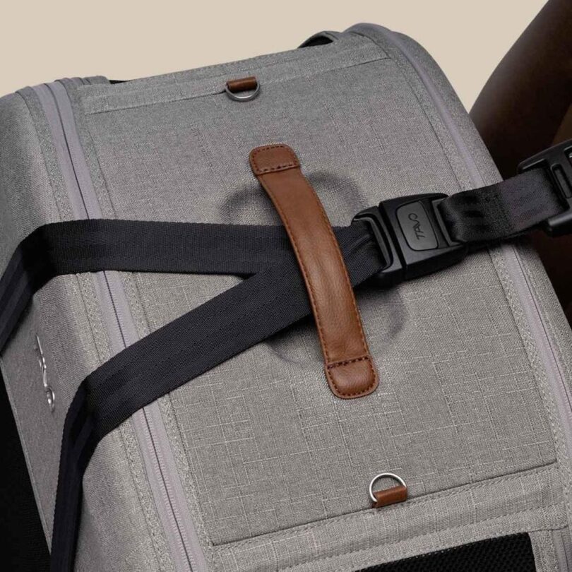 Close-up of a gray fabric pet carrier with brown leather handle and a black adjustable strap fitted through a slot